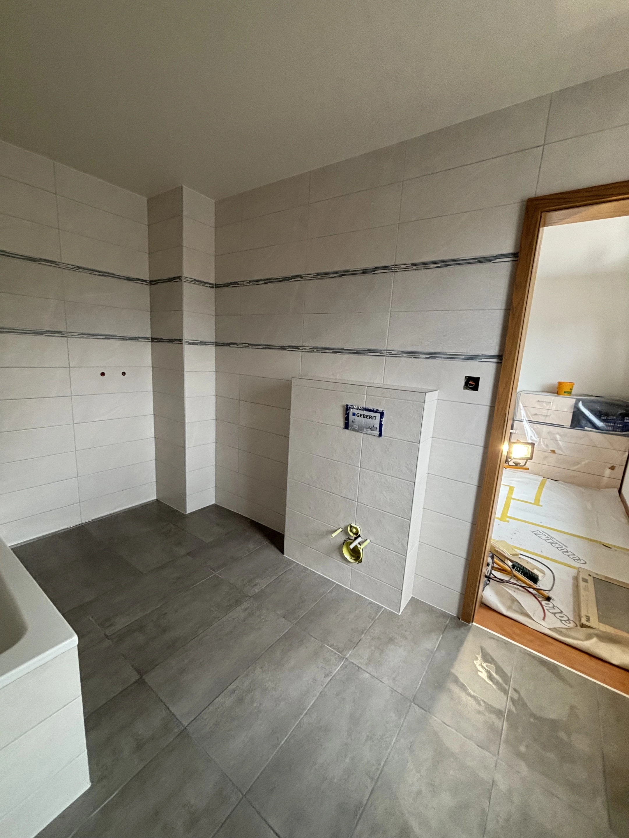 Faïence claire et frises décoratives côté entrée, avec habillage du bâti sanitaire et carrelage gris au sol